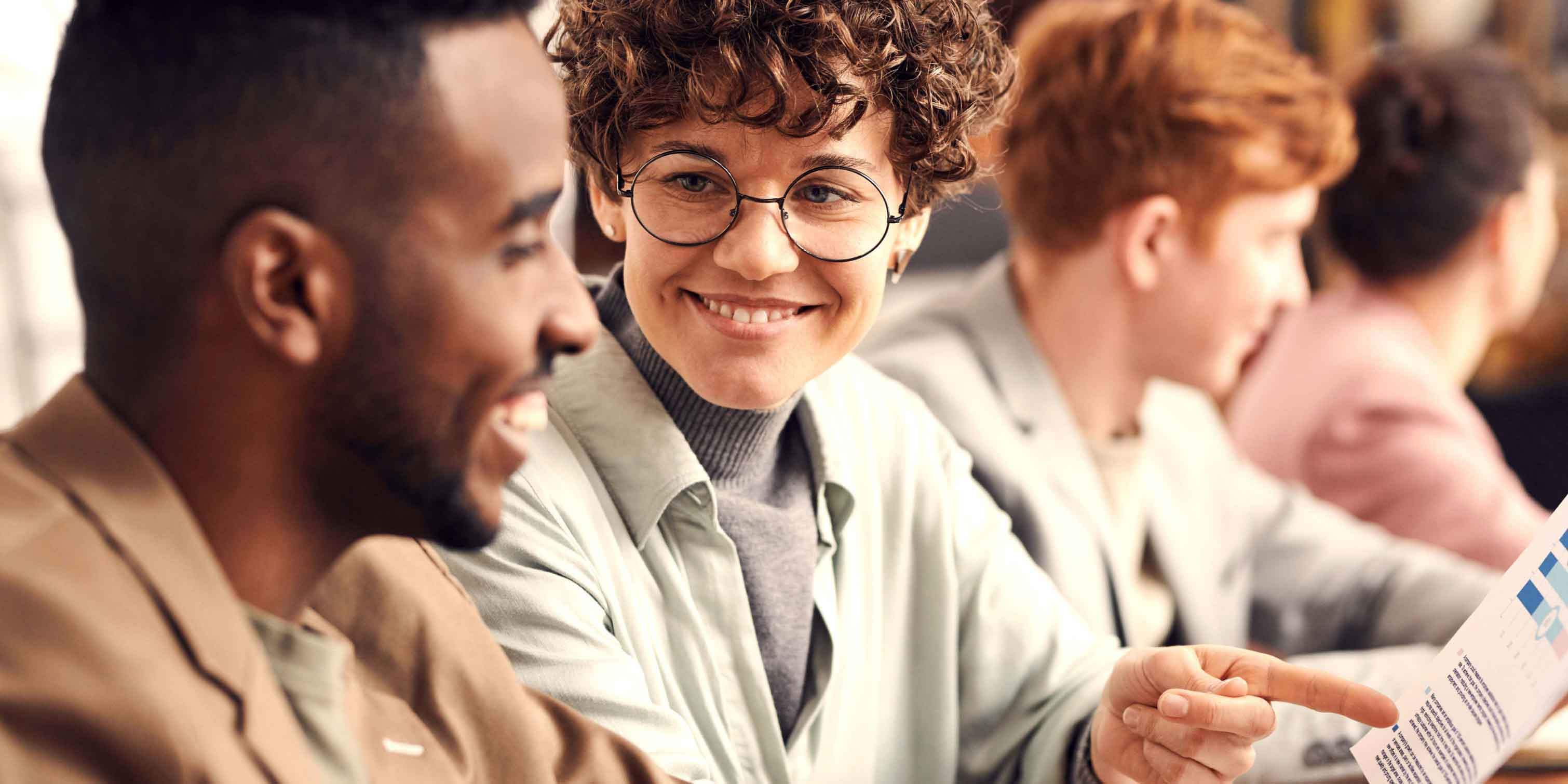 A photograph of a woman and a man at an office. They are both seated and the man is looking at a paper document. The woman is pointing at the document and is smilingly looking at the man.