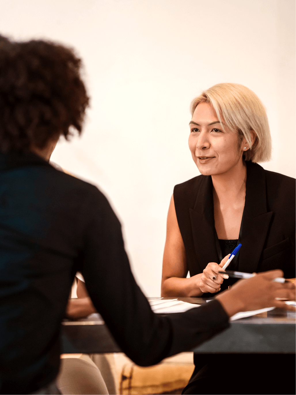 A photograph of two women sitting on opposite sides of a table in an office environment. One of the women is facing the camera looking at the women opposite her. The other woman has her back to the camera.