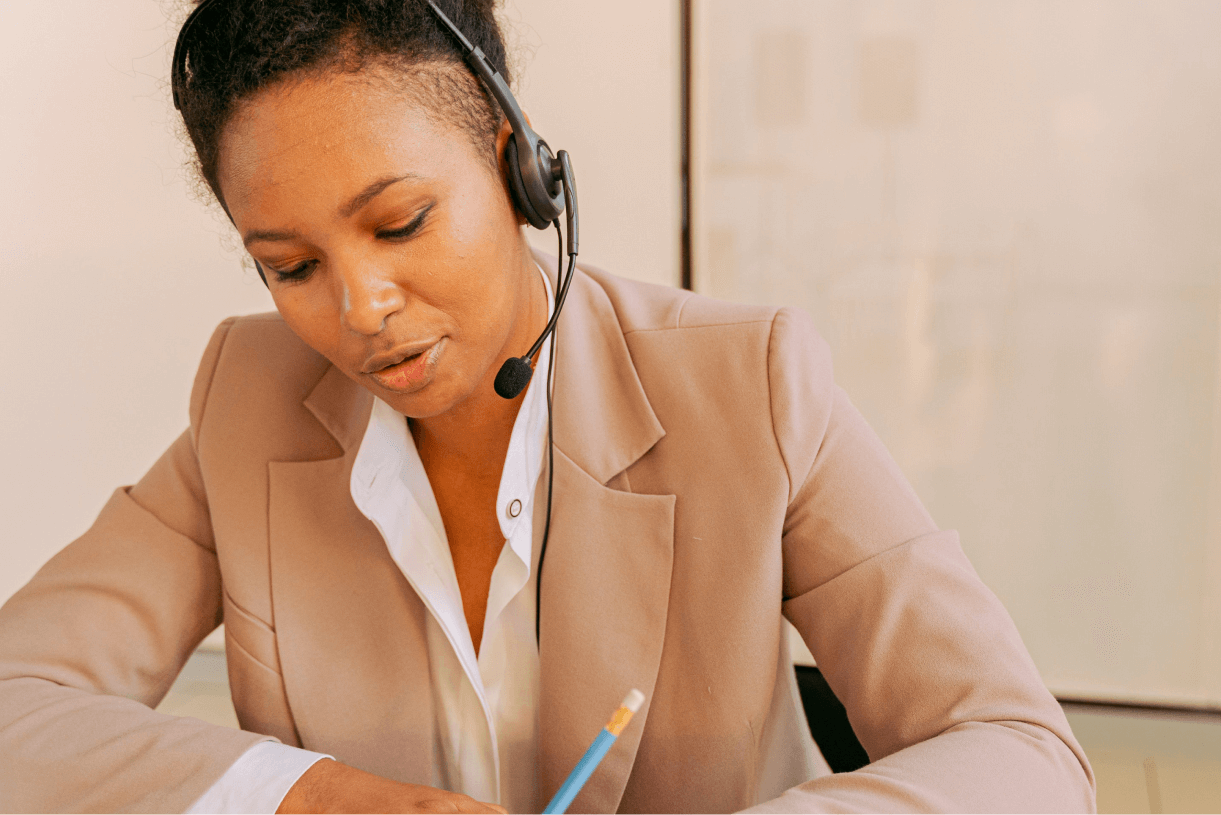 A photograph of a woman with a headset in an office environment. She is gripping a pen and gazing downwards. It appears she is taking notes.